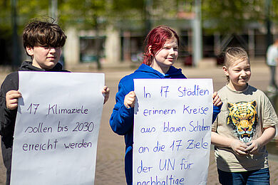 Drei Jugendliche halten Plakate hoch und lächeln. Auf einem Plakat steht "17 Klimaziele sollen bis 2030 erreicht werden", auf dem anderen "In 17 Städten erinnern Kreise aus blauen Schuhen an die 17 Ziele der UN für nachhaltige..." (der Rest ist abgeschnitten).