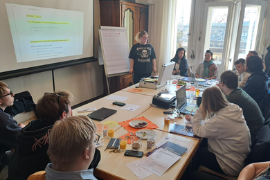 Ein großes Flipchart wird von zwei Händen gehalten. Darauf stehen handschriftlich Stichpunkte zu Themen wie Ausstattung, Kommunikation, Mitbestimmung und Ideen. Daneben sind farbige Notizzettel angebracht, auf denen Kontaktmöglichkeiten und Aufgaben notiert sind.