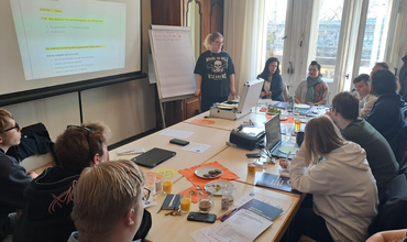 Ein großes Flipchart wird von zwei Händen gehalten. Darauf stehen handschriftlich Stichpunkte zu Themen wie Ausstattung, Kommunikation, Mitbestimmung und Ideen. Daneben sind farbige Notizzettel angebracht, auf denen Kontaktmöglichkeiten und Aufgaben notiert sind.