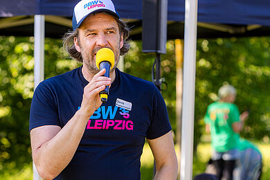 Ein Mann steht mit einem gelben Mikrofon vor einer Bühne und spricht. Er trägt ein Cap und T-Shirt, mit einer blau und pinkfarbenen Aufschrift BBW Leipzig 35.