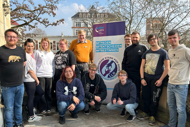 Eine Gruppe von Personen steht im Freien vor einer Mauer und Bäumen und blickt in die Kamera. Eine Person im Vordergrund zeigt ein Peace-Zeichen, während die anderen locker nebeneinander stehen. Die Gruppe wirkt entspannt und wie bei einem gemeinsamen Treffen.