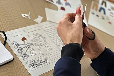Eine Person sitzt an einem Tisch und schneidet Papier mit einer Schere aus. Daneben liegt ein Arbeitsblatt mit Zeichnungen und handschriftlichen Notizen sowie weitere kreative Materialien.