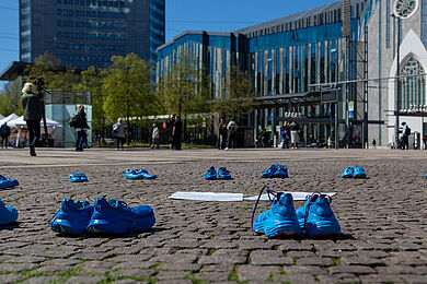 Mehrere Paare leuchtend blauer Schuhe stehen auf dem gepflasterten Boden. Im Hintergrund ist das Augusteum der Universität Leipzig zu sehen.