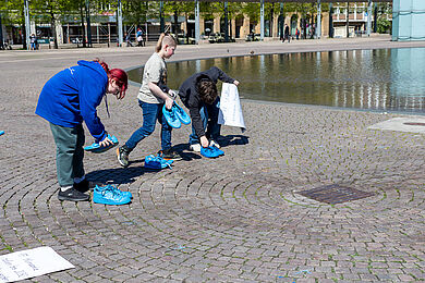Mehrere Jugendliche verteilen leuchtend blau bemalte Schuhe vor dem großen Teich auf dem Augustusplatz.