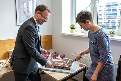 Bastian Schmiedel, rechts, und Lea Beuter, links, schneiden den Kuchen an. Der Kuchen ist mit Schokolade überzogen und ist beschriftet