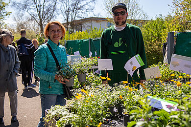 Eine Frau hält zwei Pflanztöpfe in der Hand. Neben ihr steht ein Auszublidender aus dem Fachbereich Gartenbau. Beide schauen freundlich in die Kamera. Im Vordergrund sieht man weitere Pflanztöpfe, die zum Verkauf stehen.