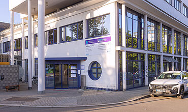 Haupteingang der Witzgallstraße 6 mit der Beschilderung für das BBW und die Diakonie am Thonberg. Der Eingang befindet sich unter einem Vordach, das von hohen Säulen getragen wird. Rechts am Haus ist eine große Fensterfront. Ein helles Auto parkt davor.