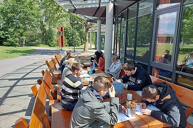 Eine Gruppe von Teilnehmenden sitzt auf den Holzbänken vorm Freizeithaus und arbeitet. Sie haben Stifte und Papier vor sich liegen.