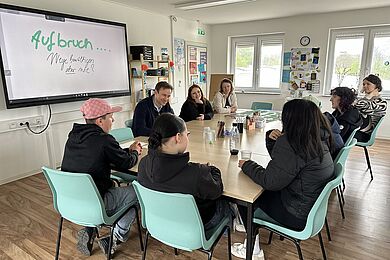 Mehrere junge Menschen und Fachkräfte sitzen gemeinsam an einem Tisch in einem Gruppenraum. Auf einem Bildschirm steht der Schriftzug „Aufbruch … Wege bewältigen aber wie?“.
