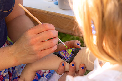 Eine Hand malt einen Schmetterling mit gelber Farbe auf einen Kinderarm.