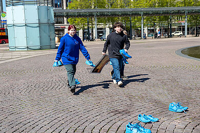Mehrere Jugendliche verteilen leuchtend blau bemalte Schuhe vor dem großen Teich auf dem Augustusplatz.