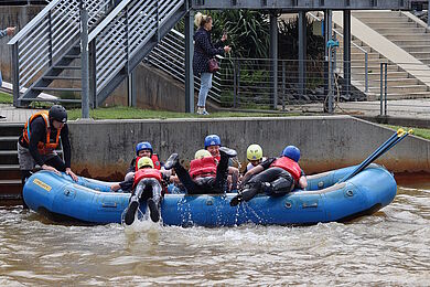 Mehrere Personen an Bord bzw. am Rande eines blauen Schlauchbootes im Wasser. Die Personen tragen Helme. Eine erwachsene Person links hält das Boot fest.