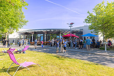 Viele Personen stehen vor einem Gebäude und unterhalten sich ausgiebig. Außerdem ist ein Sonnenschirm in rot aufgestellt, die Sonne scheint. Das Gebäude grenzt an eine schöne grüne Wiese, auf welcher im Vordergrund ein pinkfarbener Liegestuhl steht.