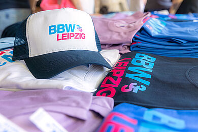 Merchandise Artikel zum 35. Jubiläum des BBW: Kappe mit Aufdruck "BBW Leipzig 35" in Hellblau und Pink, außerdem T-Shirts in verschiedenen Farben.