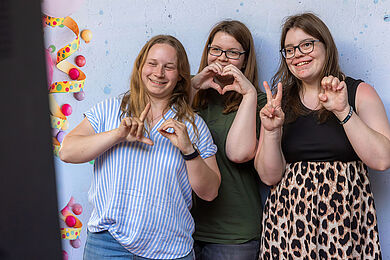 Drei junge Frauen vor einer Fotowand, formen mit ihren Händen das Wort "LOVE".