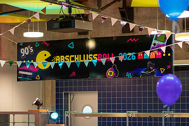 Großes Banner im Stile der 90er mit der Aufschrift "Abschlussball 2026", Davor bunte Girlanden und Luftballons