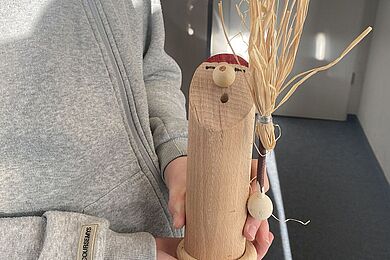 Eine Person in einer grauen Sweatshirtjacke, die man nur bis zur Brust sieht, hält eine Weihnachtsmannfigur, die aus einem runden Holzstück und anderen Naturmaterialien gefertigt wurde. 