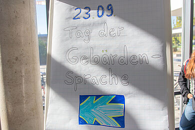 Auf einer Flipchart steht in großen Buchstaben „23.09. Tag der Gebärdensprache“. Darunter ist die Flagge der Gehörlosen- und Gebärdensprachgemeinschaft aufgemalt.