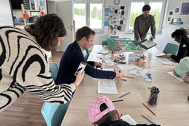 Mehrere junge Menschen und Fachkräfte sitzen gemeinsam an einem großen Tisch und bearbeiten kreative Aufgaben mit Stiften und Arbeitsblättern. Eine Person dokumentiert die Situation mit einem Smartphone.