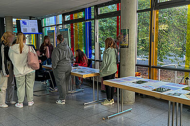Im Foyer des Berufsbildungswerks Leipzig stehen viele Menschen an einem langen Tisch mit Informationsmaterial und unterhalten sich oder machen ein Spiel, das auf dem hinteren Tisch aufgebaut ist.