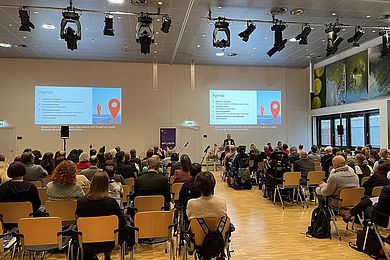 In einem großen Saal sind viele sitzende Menschen zu sehen, auf der Bühne hält Prof. Böhm einen Vortrag bei der Inklusionskonferenz. Die Agenda ist durch einen Beamer an die Wand hinter ihm angezeigt.