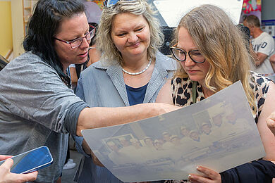 Drei Frauen stehen in einem Raum und schauen sich gemeinsam ein großes Foto an, was eine Frau von ihnen in der Hand hält. 