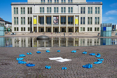 Mehrere Paare leuchtend blauer Schuhe stehen auf dem gepflasterten Boden. Im Hintergrund ist die Leipziger Oper zu sehen.