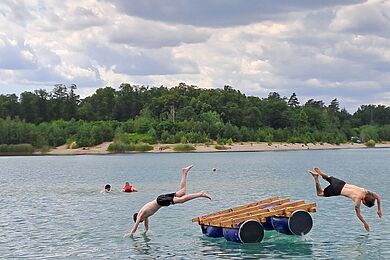 Blick über einen See bis zu einer Insel oder einem anderen Ufer. Im Vordergrund springen zwei Menschen von einem selbstgebauten Floß per Kopfsprung ins Wasser.