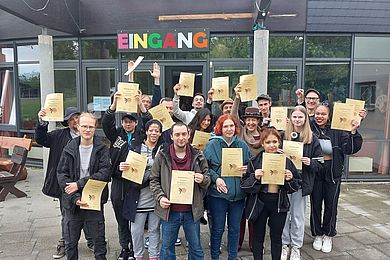Eine Gruppe von Teilnehmenden präsentiert stolz ihre Urkunde über die Teilnahme am Theatertreffen. Im Hintergrund ist in bunten Buchstaben "Eingang" zu lesen (es handelt sich um den Eingang zum Freizeithaus).