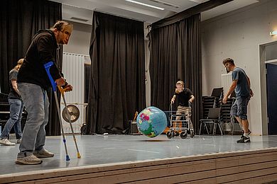 Performance auf der Bühne bei der Theatershow - vier Personen spielen Theater. Sie haben Masken auf. Eine Person vorne links läuft mit Krücken. In der Mitte dotzt ein Wasserball bedruckt mit der Erdkügel auf. 