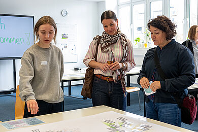 Eine junge Frau in einem grauen Sweatshirt erklärt zwei weiteren Frauen etwas auf einem Tisch