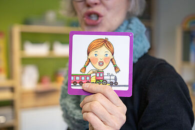 Logopädin hält ein Bild mit dem "Mundbild SCH". Man sieht, wie sie im Hintergrund das Mundbild formt.