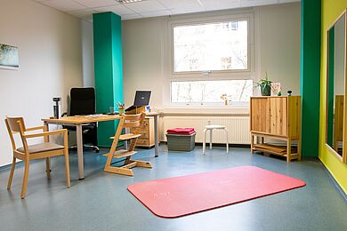 Therapieraum mit Tisch, Stühlen und Kinderstuhl, auf dem Boden eine rote Matte, rechts ein Spiegel an der Wand