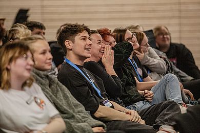 Blick ins Publikum bei der inklusiven Abendshow. Die Gäste lachen. Im Fokus ist ein lachender junger Mann mit kurzen dunklen Haaren, er trägt ein schwarzes Hemd und hat die Ärmel hochgekrempelt. Um den Hals hat er ein blaues Schlüsselband mit einer Karte dran.