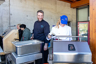 Ausbilder und Auszubildende am Essensstand. Sie stehen hinter großen Edelstahl-Warhmhaltebehältern