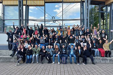 Alle Teilnehmenden auf der Steintribüne vor der Sporthalle. Die Personen winken in die Kamera.