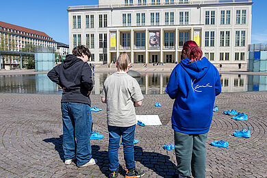 Drei Jugendliche stehen vor einem Kreis aus leuchten blau bemalten Schuhpaaren, die auf dem Augustusplatz aufgereiht stehen. Sie blicken hinab, in der Mitte des Kreises ist ein Plakat platziert. Im Hintergrund ist die Leipziger Oper zu sehen.