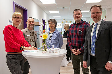Fünf Personen hinter einem Stehtisch mit einem Blumenstrauß. Alle schauen lächelnd in die Kamera.