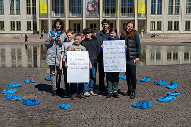 Eine Gruppe erwachsener Personen und Jugendlicher steht in einem großen Kreis aus leuchtend blau angemalten Schuhen. Sie lächeln in die Kamera und halten zwei Plakate in die Höhe.