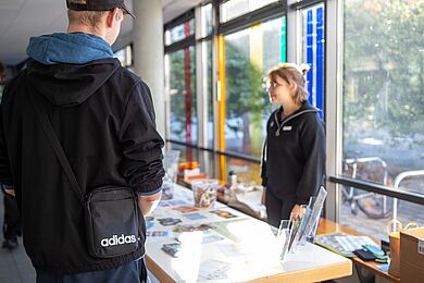 Eine Person ist scharf im Vordergrund von hinten zu sehen. Sie steht an einem Informationstisch mit verschiedenen Flyern und Informationsblättern. Hinter dem Tisch im Foyer des Berufsbildungswerks Leipzig steht eine*r Mitarbeiter*in, unscharf zu sehen.